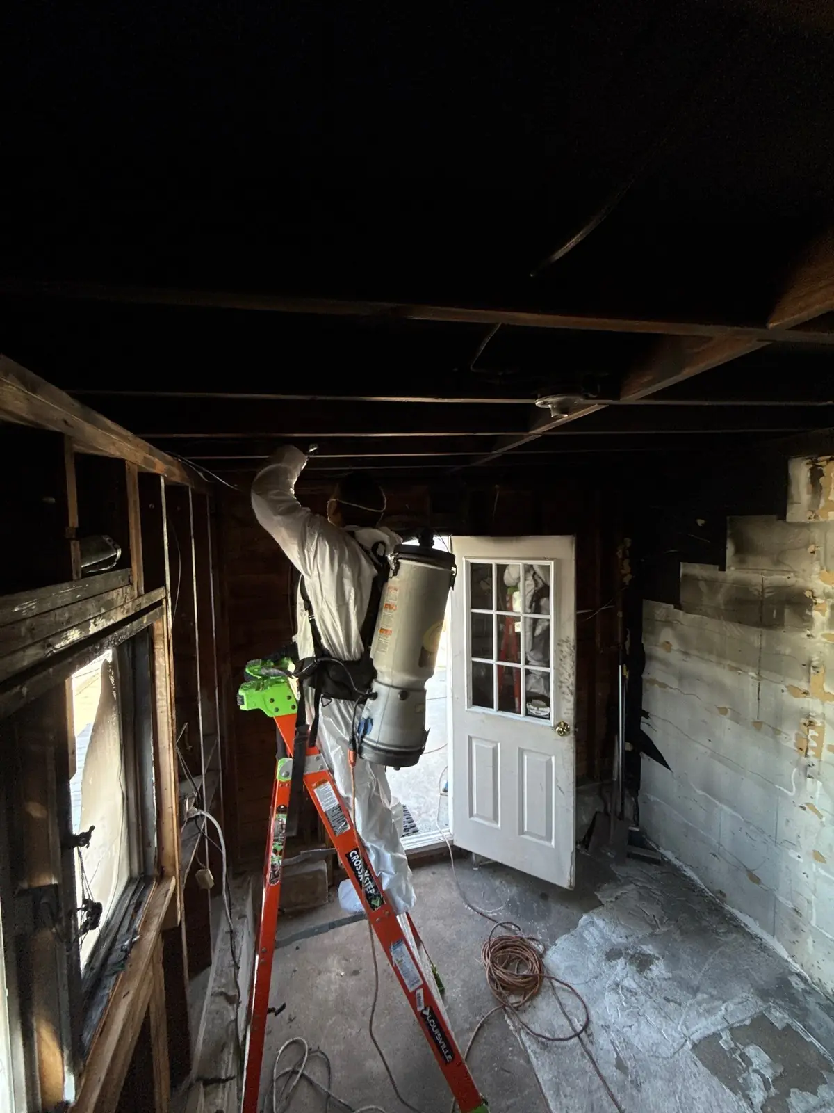 K.O.S. technician in PPE on a ladder, HEPA-vacuuming soot from the ceiling with a backpack vacuum