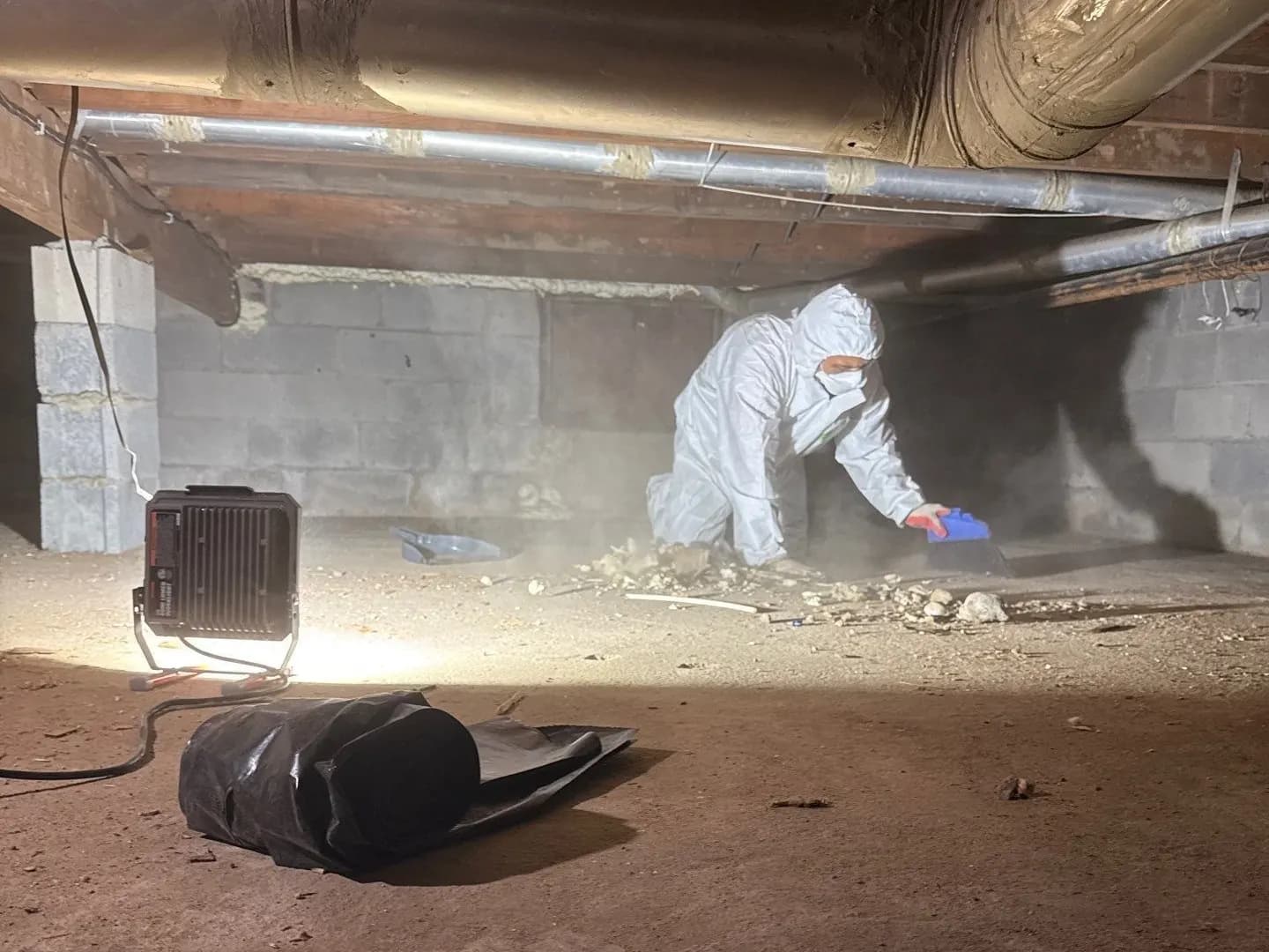 K.O.S. handler in a protective suit and respirator removing asbestos debris in a contained crawlspace