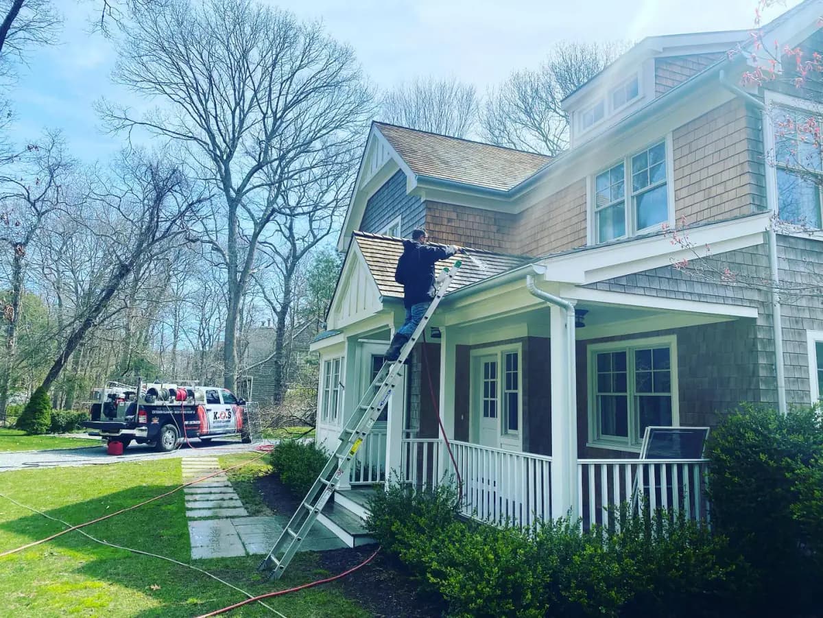 K.O.S. technician on a ladder at the porch roofline of a Long Island home, the company truck in the driveway