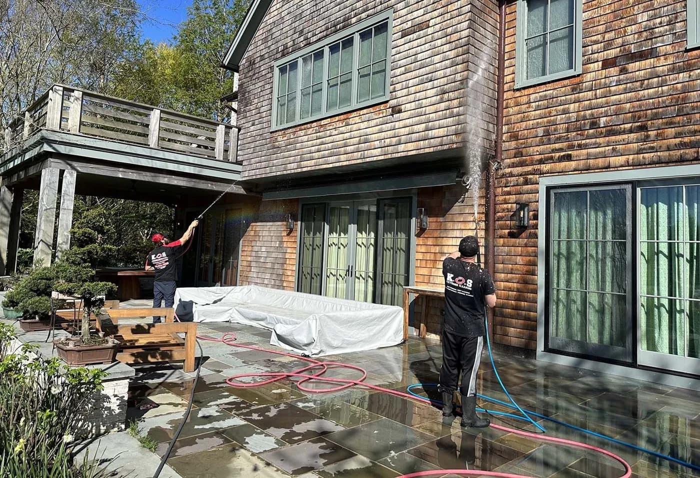 Two K.O.S. technicians soft-washing cedar siding from a stone patio, furniture covered